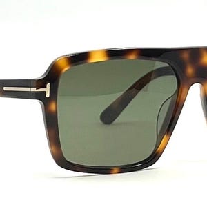 Może przedstawiać: Okulary przeciwsłoneczne w wzór żółwia z ciemnozielonymi soczewkami. Okulary mają kwadratową oprawkę i srebrny akcent na zausznikach. Nazwa marki jest widoczna na prawym zauszniku.