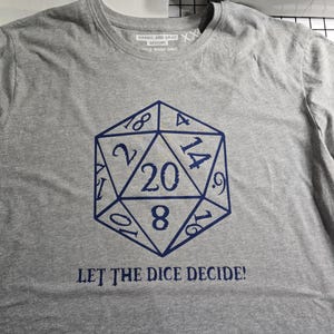 May include: A grey t-shirt with a blue twenty-sided die graphic, displaying the numbers 2, 8, 9, 10, 14, 16, and 18. The text "LET THE DICE DECIDE!" is below the die. The shirt also has the text "Harris and Gray Designs".