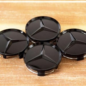 May include: Four black wheel center caps with the Mercedes-Benz logo. Each cap features a glossy black finish and a three-pointed star emblem. The caps are circular with a silver retaining ring.