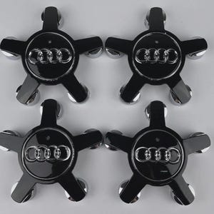 May include: Four black and silver car wheel center caps. Each cap has a five-point star design with a silver Audi logo in the center. The caps have a glossy finish and chrome-colored accents.