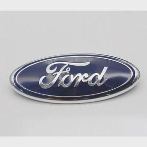 Puede incluir: Un emblema ovalado de Ford con un fondo azul oscuro y un borde plateado. La palabra "Ford" está escrita en una fuente plateada estilizada en el centro del emblema.