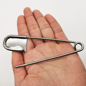 May include: A large, silver-colored safety pin. The pin is held in a hand, showcasing its size and design. The pin has a rounded head and a sharp point, typical of safety pins used for various purposes.