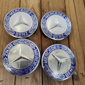May include: Four silver and blue Mercedes-Benz hubcaps. Each hubcap features the Mercedes-Benz logo, a three-pointed star, in the center, surrounded by a blue ring with the words "Mercedes" and "Benz". The hubcaps are on a wooden surface.