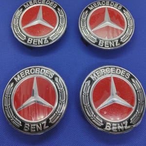 May include: Four Mercedes-Benz wheel center caps. Each cap features a red center with a silver three-pointed star emblem, surrounded by a silver ring and the words "MERCEDES BENZ" in black lettering. The caps are on a blue background.