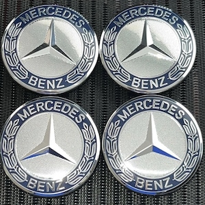 Op de afbeelding: Vier zilver-blauwe Mercedes-Benz wieldopjes. Elke dop heeft het Mercedes-Benz-logo, een driepuntige ster, omringd door een laurierkrans en het woord "Mercedes" boven "Benz".
