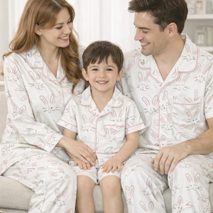May include: Family pyjamas in white with a repeating bunny face design and pink trim. The set includes long-sleeved shirts and trousers for adults, and a short-sleeved shirt and shorts for the child.