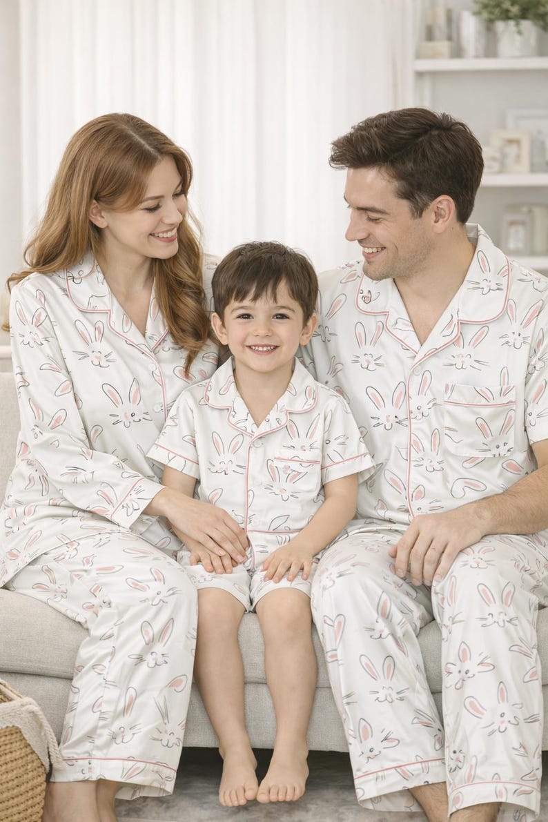 May include: Family pyjamas in white with a repeating bunny face design and pink trim. The set includes long-sleeved shirts and trousers for adults, and a short-sleeved shirt and shorts for the child.