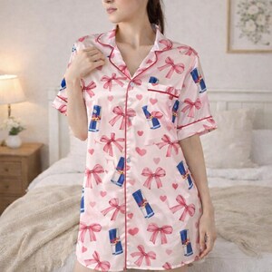 May include: Pink satin pajama shirt dress with a pattern of red bows, blue and white energy drink cans, and pink hearts. The dress has short sleeves, a collared neckline, and red trim.