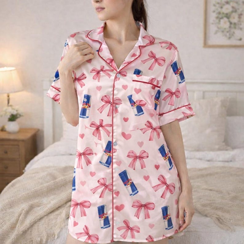 May include: Pink satin pajama shirt dress with a pattern of red bows, blue and white energy drink cans, and pink hearts. The dress has short sleeves, a collared neckline, and red trim.
