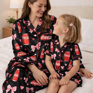 K&ouml;nnte beinhalten: Schwarzer Coca-Cola-Pyjama mit roter Paspel, verziert mit Coca-Cola-Logos, rosa Schleifen und Herzen. Eine Frau und ein Kind tragen den Pyjama auf einem Bett. Der Pyjama ist aus einem weichen, seidigen Material.