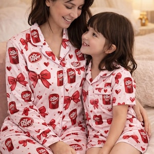 May include: Matching pink pajamas with a Dr. Pepper soda can and red bow pattern. The pajamas are worn by two people. The long-sleeved top and pants are worn by one person, and the short-sleeved top and shorts are worn by the other.