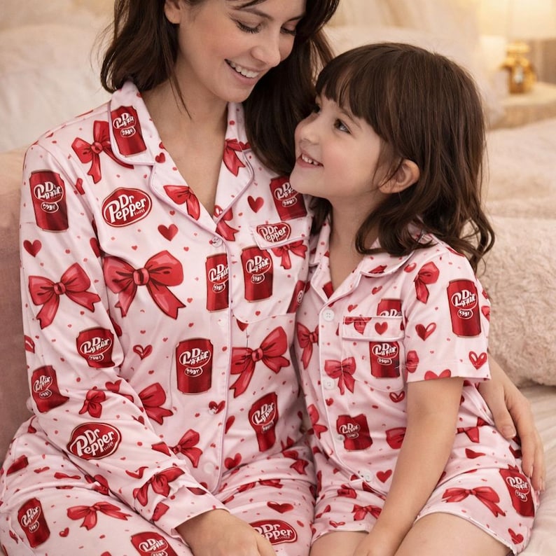 May include: Matching pink pajamas with a Dr. Pepper soda can and red bow pattern. The pajamas are worn by two people. The long-sleeved top and pants are worn by one person, and the short-sleeved top and shorts are worn by the other.