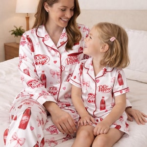 May include: Matching white Dr. Pepper pajamas with red trim, featuring Dr. Pepper bottles and bows. A woman and a child are wearing the pajamas while sitting on a bed. The pajamas have the Dr. Pepper logo.