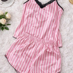 Könnte beinhalten: Rosa-weiß gestreiftes Pyjama-Set. Das Camisole-Top hat schwarze Spitzentrimm und dünne Träger. Die Shorts haben einen elastischen Bund und schwarze Paspeln. Das Set wird auf einer weißen, flauschigen Oberfläche präsentiert. Der Text "MirageShopDesign" befindet sich unten.