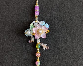 May include: A floral keychain with a gold-colored heart-shaped clasp. The charm features a pink flower with a pearl center, surrounded by blue and clear flowers, and colorful beads. The keychain is approximately 5 inches long.