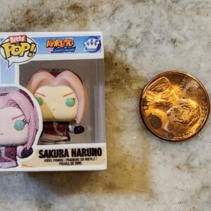 May include: A small, boxed Sakura Haruno vinyl figure from the Naruto anime series. The figure has pink hair and green eyes. A penny is next to the figure for size comparison.