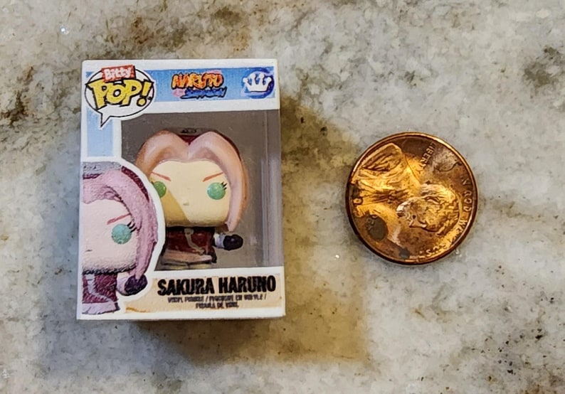 May include: A small, boxed Sakura Haruno vinyl figure from the Naruto anime series. The figure has pink hair and green eyes. A penny is next to the figure for size comparison.