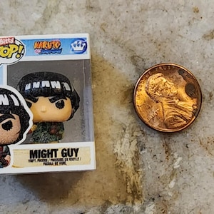 May include: A Funko Pop! vinyl figure of Might Guy from the Naruto anime series, displayed in its original packaging. A US penny is next to the figure for size comparison. The figure has a black bowl cut hairstyle and a green outfit.