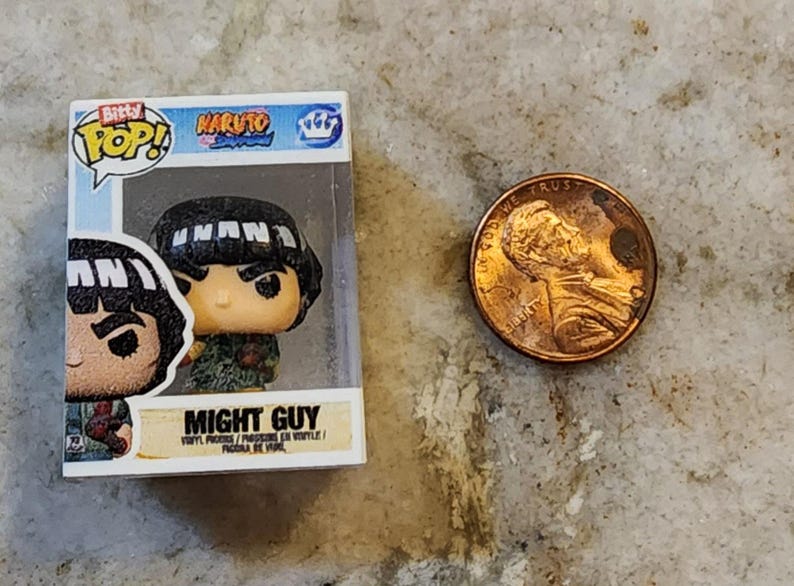 May include: A Funko Pop! vinyl figure of Might Guy from the Naruto anime series, displayed in its original packaging. A US penny is next to the figure for size comparison. The figure has a black bowl cut hairstyle and a green outfit.