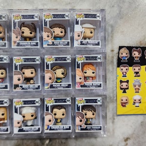 May include: A set of 12 Friends TV show character figurines in clear plastic display cases. The figurines are of Rachel, Chandler, Ross, Joey, Monica, Phoebe, and the characters are holding various props from the show. The figurines are all in the same style and pose, and they are all wearing their signature outfits from the show.