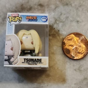 May include: A small Tsunade vinyl figure from the Naruto anime series, packaged in a clear plastic box. The figure has blonde hair and is in a seated position. A penny is next to the figure for size comparison.