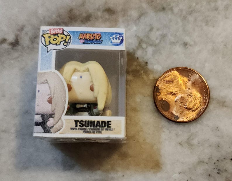 May include: A small Tsunade vinyl figure from the Naruto anime series, packaged in a clear plastic box. The figure has blonde hair and is in a seated position. A penny is next to the figure for size comparison.