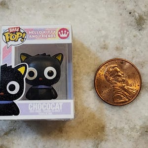 Puede incluir: Una figura en miniatura de Chococat de la serie Hello Kitty and Friends, exhibida en una caja de pl&aacute;stico transparente. La figura del gato negro tiene ojos blancos grandes, puntas de orejas amarillas y un collar morado. Una moneda est&aacute; al lado de la figura.