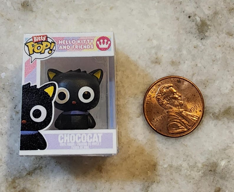 Puede incluir: Una figura en miniatura de Chococat de la serie Hello Kitty and Friends, exhibida en una caja de pl&aacute;stico transparente. La figura del gato negro tiene ojos blancos grandes, puntas de orejas amarillas y un collar morado. Una moneda est&aacute; al lado de la figura.