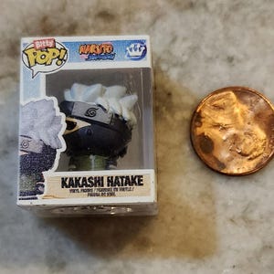 May include: A miniature Kakashi Hatake vinyl figure from the Naruto Shippuden series, in a clear plastic display case. The figure has white hair, a black mask, and a green outfit. A penny is next to the figure for size comparison.