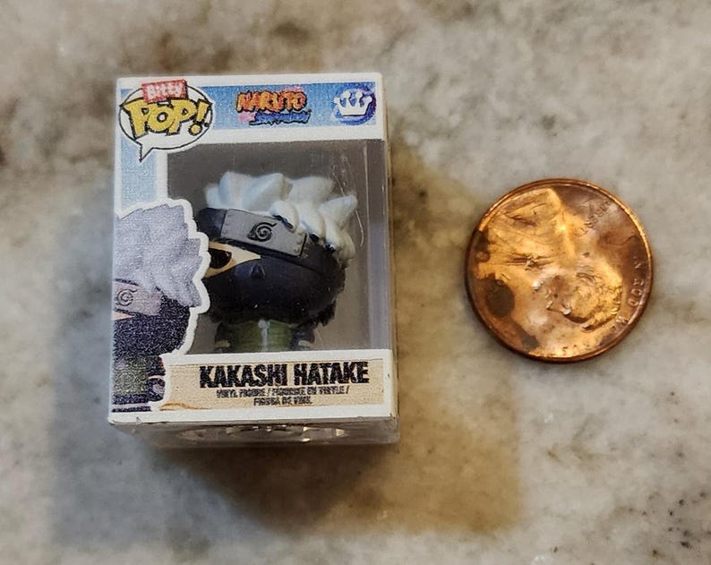 May include: A miniature Kakashi Hatake vinyl figure from the Naruto Shippuden series, in a clear plastic display case. The figure has white hair, a black mask, and a green outfit. A penny is next to the figure for size comparison.