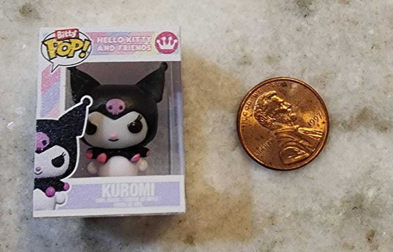 Puede incluir: Una figura de Bitty Pop! de Kuromi de Hello Kitty and Friends, en una vitrina de pl&aacute;stico transparente. La figura es negra, blanca y rosa. Una moneda de un centavo estadounidense est&aacute; al lado de la figura.
