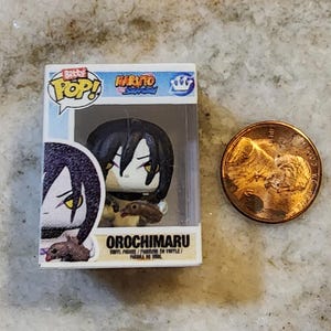 May include: A miniature Orochimaru vinyl figure from the Naruto anime series, displayed in its original box. The figure has black hair and yellow eyes. A penny is next to the figure for size comparison. The box reads "Bitty Pop!" and "Orochimaru".