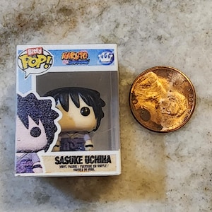 May include: A Funko Pop! vinyl figure of Sasuke Uchiha from the Naruto anime series, displayed in its original box. The figure has black hair and is wearing a purple outfit. A penny is next to the figure for size comparison.