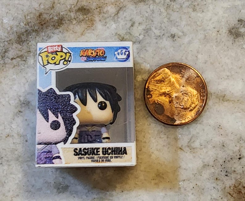 May include: A Funko Pop! vinyl figure of Sasuke Uchiha from the Naruto anime series, displayed in its original box. The figure has black hair and is wearing a purple outfit. A penny is next to the figure for size comparison.