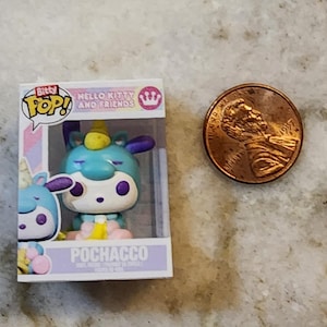 Puede incluir: Una figura de Pochacco de Hello Kitty and Friends en una caja de exhibici&oacute;n, junto a un centavo. La figura es un unicornio azul claro con un cuerno amarillo y orejas moradas. La caja tiene el logo de Bitty Pop!