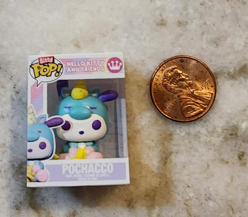 Puede incluir: Una figura de Pochacco de Hello Kitty and Friends en una caja de exhibici&oacute;n, junto a un centavo. La figura es un unicornio azul claro con un cuerno amarillo y orejas moradas. La caja tiene el logo de Bitty Pop!