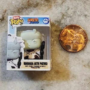 May include: A small, boxed "Bitty Pop!" figure of Madara (6th Paths) from the Naruto series. The figure is in a clear plastic case next to a US penny. The box is white with blue and yellow accents. The figure is primarily green, white, and black.