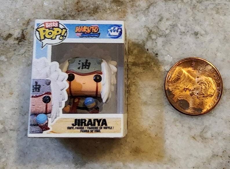 May include: A miniature Jiraiya vinyl figure from the Naruto anime series, displayed in a clear plastic box. The figure has white hair, a green headband, and a brown outfit. A penny is next to the figure for size comparison.