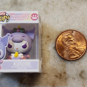 Puede incluir: Una figura Bitty Pop! Hello Kitty and Friends de Kuromi en una vitrina de pl&aacute;stico transparente. La figura es morada, blanca y amarilla. Una moneda est&aacute; al lado de la figura para comparar el tama&ntilde;o.