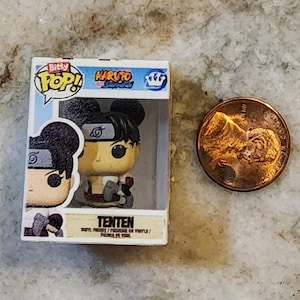 May include: A small, boxed "Bitty Pop!" figure of Tenten from Naruto, next to a penny. The figure is in a clear plastic case with the "Naruto" and "Bitty Pop!" logos. The figure is tan, black, and red. The penny is copper-colored.