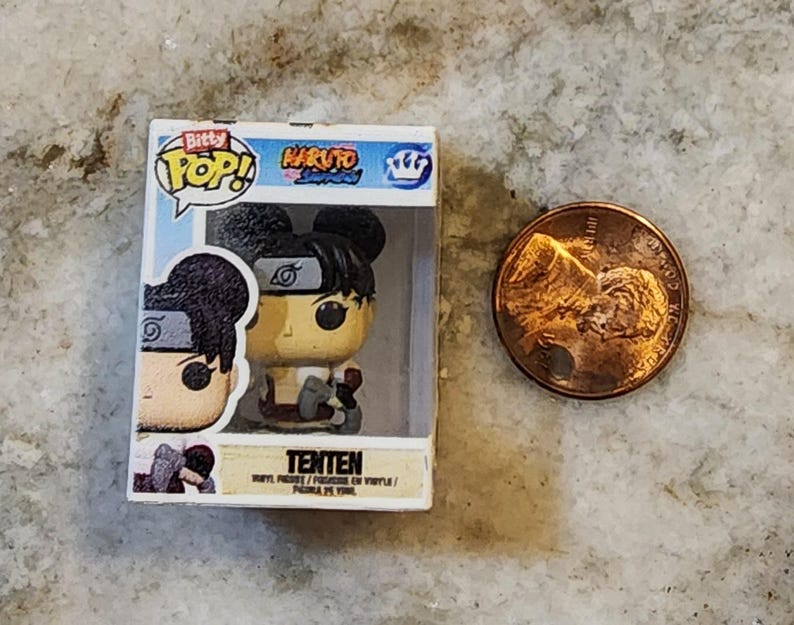 May include: A small, boxed "Bitty Pop!" figure of Tenten from Naruto, next to a penny. The figure is in a clear plastic case with the "Naruto" and "Bitty Pop!" logos. The figure is tan, black, and red. The penny is copper-colored.