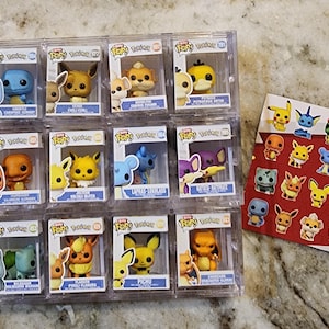 May include: A collection of Funko Pop! figures of various Pokémon characters in clear display cases. A penny is next to the figures for scale. A red card with Pokémon stickers is also in the image.
