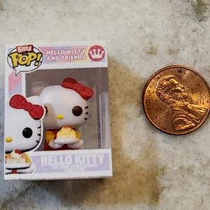 Puede incluir: Una figura de Bitty Pop! de Hello Kitty and Friends en una caja de exhibici&oacute;n, junto a un centavo estadounidense. La figura es blanca con un lazo rojo y sostiene un peque&ntilde;o pastel. La caja dice "Hello Kitty and Friends".