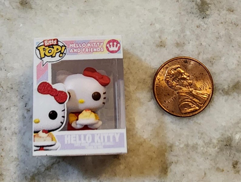 Puede incluir: Una figura de Bitty Pop! de Hello Kitty and Friends en una caja de exhibici&oacute;n, junto a un centavo estadounidense. La figura es blanca con un lazo rojo y sostiene un peque&ntilde;o pastel. La caja dice "Hello Kitty and Friends".