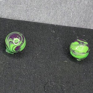 May include: Two round, marbled beads. One bead is green and purple with a swirl pattern. The other bead is green with a swirl pattern. The beads are on a black surface.