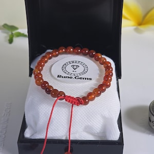 May include: A beaded bracelet with reddish-brown beads, presented in a black box with a white interior. The bracelet is secured with a red string and a knot. The box contains a white label with the text "Rune.Gems".