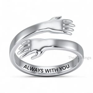 May include: A silver-colored ring with a design of two hands embracing. The ring is engraved with the words "ALWAYS WITH YOU". The ring is a piece of jewelry.
