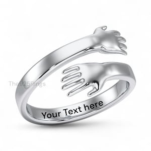 May include: A silver-colored ring with a hugging hands design. The ring features two hands that appear to be embracing. The words "Your Text here" are engraved on the inside of the ring.
