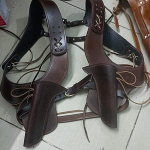 Op de afbeelding: Een bruin leren pistoolholster-rig met twee holsters, schouderbanden en riemlussen. De rig heeft decoratieve stiksels en messing gespen. Het leer lijkt goed gemaakt en duurzaam, geschikt voor historische re-enactments of kostuums.