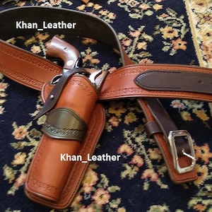 Puede incluir: Un juego de cinturón y funda de cuero marrón con una hebilla plateada. La funda contiene un revólver. El cinturón y la funda están sobre una alfombra estampada. La funda tiene una placa decorativa de metal.